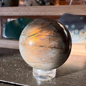 May include: A polished, iridescent sphere, likely a gemstone or mineral specimen, rests on a clear acrylic stand. The sphere displays a mix of brown, gold, and gray hues with a smooth, reflective surface. The overall theme is natural decor.