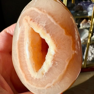 May include: A polished, egg-shaped agate geode. The geode features concentric bands of peach, cream, and brown hues, with a hollow center revealing a crystal-lined cavity. The stone is held in a hand.