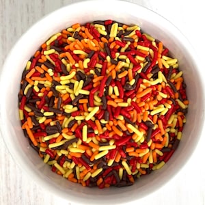 May include: A white bowl filled with colorful sprinkles. The sprinkles are a mix of red, orange, yellow, and brown, creating a vibrant and appetizing display. The sprinkles are small, elongated shapes, perfect for decorating baked goods.