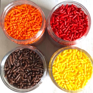 May include: Four clear jars filled with colorful sprinkles. The sprinkles are orange, red, brown, and yellow. The jars are arranged in a square formation, perfect for baking and decorating desserts.