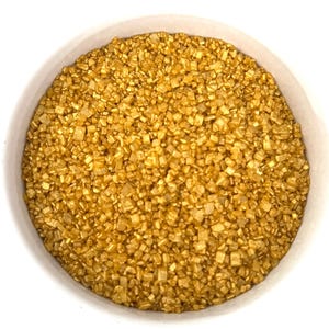 May include: A close-up of a white bowl filled with gold sugar sprinkles. The sprinkles are small and have a shiny, metallic finish.