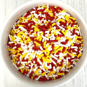 May include: A white ceramic bowl filled with colorful sprinkles. The sprinkles are a mix of red, yellow, and white, with a rectangular shape. The bowl is set against a white wooden surface.