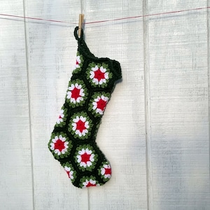May include: A handmade Christmas stocking, crocheted with a dark green yarn. The stocking features a pattern of red and white flower-like shapes. It is hanging from a red string with a wooden clothespin against a white wooden background.