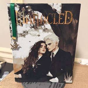 May include: A hardback book titled "Mangled" with a gold foil title and a bookmark. The cover art depicts a couple in formal attire, set against a dark backdrop. A green ribbon bookmark with a star charm is attached.