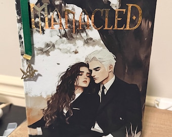 May include: A hardcover book titled "Mangled" with a gold foil title and a bookmark. The cover art features a couple in formal attire, with a dark background. A green ribbon bookmark with a star charm is attached.