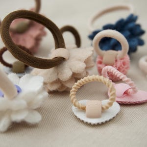 Op de afbeelding: Een verzameling haaraccessoires, waaronder met stof bedekte elastische haarbanden en decoratieve accenten in bloemvorm. Het kleurenpalet omvat tinten bruin, roze, wit en blauw. Deze haarbanden zijn ontworpen voor verschillende kapsels.