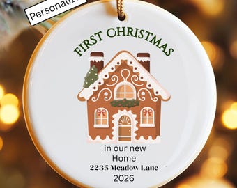 May include: A round white ceramic ornament featuring a gingerbread house illustration with green text reading FIRST CHRISTMAS and black text reading in our new Home 2235 Meadow Lane 2026. A gold string is attached to the top for hanging.