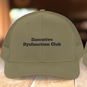 May include: An olive green trucker hat featuring the text "Executive Dysfunction Club" in dark blue. The hat has a mesh back and a curved peak. The hat is shown from the front and back.