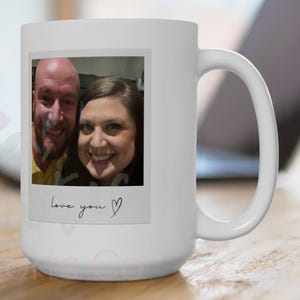 Può includere: Tazza in ceramica bianca con una fotografia stampata di una coppia sorridente. La foto è incorniciata con un bordo bianco e include il testo scritto a mano "love you" con un simbolo a forma di cuore. La tazza è su una superficie di legno.