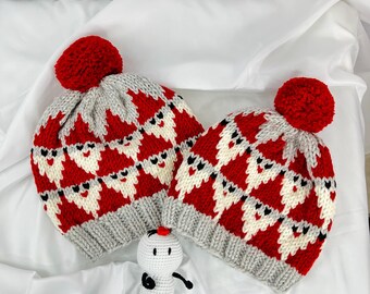 May include: Two knitted Santa hats with red bobbles and a matching snowman toy. The hats feature a repeating pattern of Santa faces in red, white, and grey. The snowman toy is white with black details and a small red hat.