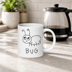 May include: White ceramic mug featuring a whimsical black line drawing of a cartoon bug with the word "BUG" beneath it. The mug is set on a countertop with a coffee maker and a plant in the background.
