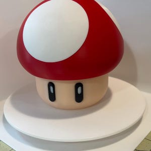 May include: A red and white mushroom-shaped decorative item. The mushroom has a red cap with a large white circle and a beige stem with black oval eyes. It is sitting on a white, round platform.