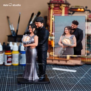 May include: A miniature wedding figurine of a couple embracing, the woman in a silver gown holding a bouquet. The man is in a black suit. The figurine is on a black base. The background includes paint bottles and brushes.