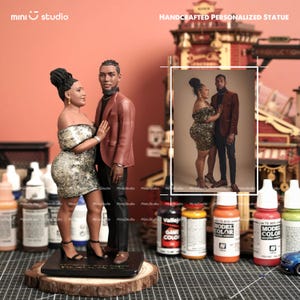 Puede incluir: Estatua personalizada y artesanal de una pareja. La mujer lleva un vestido dorado con lentejuelas y el hombre una chaqueta color óxido. La estatua está montada sobre una base de madera e incluye una foto enmarcada de la pareja.