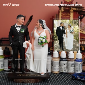 May include: A handcrafted personalized statue of a bride and groom in wedding attire. The groom wears a black tuxedo, and the bride is in a white wedding dress, holding a bouquet. The statue is on a wooden base, with a framed photo of the couple.