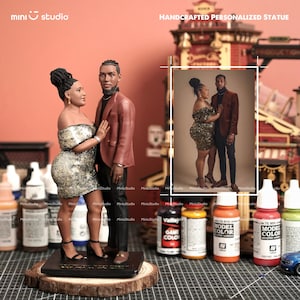 May include: A handcrafted personalized statue of a couple, featuring a woman in a gold sequined off-the-shoulder dress and a man in a rust-colored jacket. The statue is mounted on a wooden base and includes a framed photo of the couple.