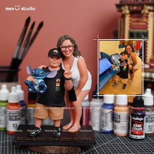 May include: A custom figurine of a child holding a Stitch plush toy, with a woman standing behind them. The figurine is on a wooden base with text. Paint bottles and brushes are in the background. The image includes a smaller photo.