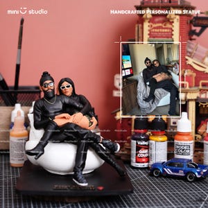 Può includere: Una statua personalizzata fatta a mano raffigurante una coppia in abiti neri, seduta su una base bianca. L'immagine include un'auto giocattolo blu in miniatura e flaconi di vernice. Il testo "mini studio" e "HANDCRAFTED PERSONALIZED STATUE" sono visibili.