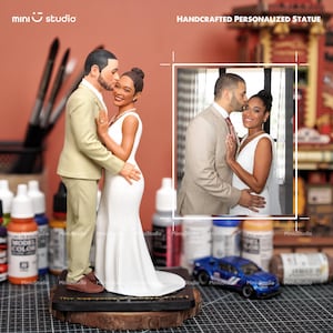 May include: A handcrafted personalized statue of a couple in wedding attire. The groom, in a tan suit, kisses the bride's forehead. The bride wears a white dress. The statue is on a wooden base. The image includes the text "mini studio" and "HANDCRAFTED PERSONALIZED STATUE".