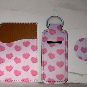 May include: Three pink and brown phone cases with a pink heart pattern. The cases are for a phone, a hand sanitizer, and a pop socket.