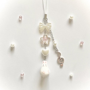Könnte beinhalten: Ein weißer Handy-Anhänger mit Schleife, Blume, Herz und Hasen-Anhänger. Der Anhänger hat eine silberne Kette mit einem Violinschlüssel und Perlenakzenten. Rosa und weiße Perlen sind um den Anhänger verstreut.
