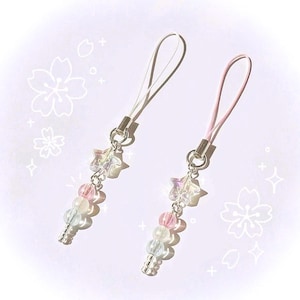 May include: Two beaded phone charms. One has a white strap, the other a pink strap. Each charm features a star-shaped bead, pink, white, and clear beads, and a silver-colored metal base. The background is white with floral and star outlines.