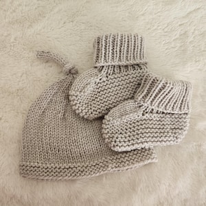 May include: A light gray knitted baby set, including a beanie hat with a knotted top and a pair of booties. The set is made of a soft, textured yarn and is displayed on a fluffy, white surface. The booties have a folded-over cuff.