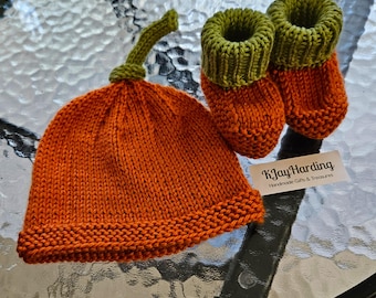 May include: A hand-knitted orange hat with a green stem top and a pair of matching orange booties with green ribbed cuffs. A small white rectangular tag with the black printed text KJay Harding Handmade Gifts & Treasures is included.