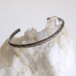 May include: A silver bangle bracelet with a decorative etched design. The bracelet is displayed on a white rock.
