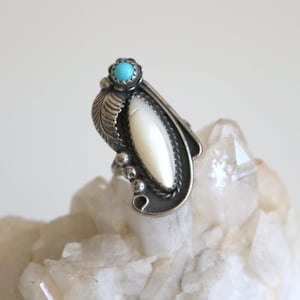 May include: A silver ring with a white mother of pearl stone and a turquoise stone. The ring is set on a bed of white crystals.