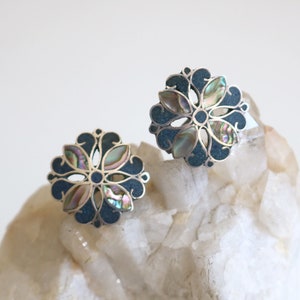 May include: A pair of silver earrings with a blue and iridescent abalone shell design. The earrings are in the shape of a flower with a circular center.