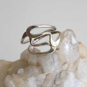 May include: A silver-toned ring featuring an open, fluid, and abstract design with interconnected curved lines. The metal band has a smooth, polished finish and a continuous, looping structure that creates negative space throughout the piece.
