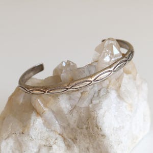 May include: A silver cuff bracelet with an open design, featuring an engraved pattern of oval shapes. The bracelet rests on a rough, white crystal formation, set against a plain white background. The bracelet's design is symmetrical.