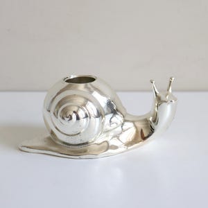 May include: A silver-colored snail-shaped decorative object. The snail has a spiraled shell and a small opening on top. The snail's body is smooth and reflective, with two small antennae. The object is likely a small vase or container.