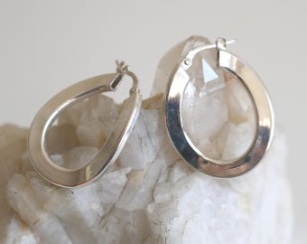 May include: A pair of silver-toned oval hoop earrings. The earrings are displayed on a white, textured surface. The earrings have a simple, polished design, with a hinged closure.