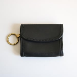 May include: A small black leather pouch with a rounded flap closure and a gold-toned metal key ring attached to the side. The accessory features visible stitching along the edges of the flap and the main body, providing a structured appearance.