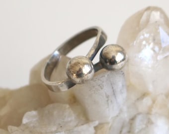 May include: A silver ring with two round balls on the top. The ring is sitting on a white crystal.