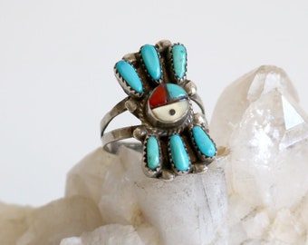 May include: A silver ring with a turquoise, red, and white sunface design. The ring features multiple turquoise stones and a central sunface with a white background and red and black accents. The ring is displayed on a clear crystal.