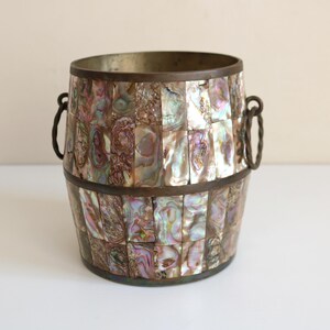 May include: A decorative barrel-shaped container with a mosaic of iridescent abalone shell pieces. The container has a metal rim and handles, and it sits on a white surface. The colors include shades of purple, green, and brown.