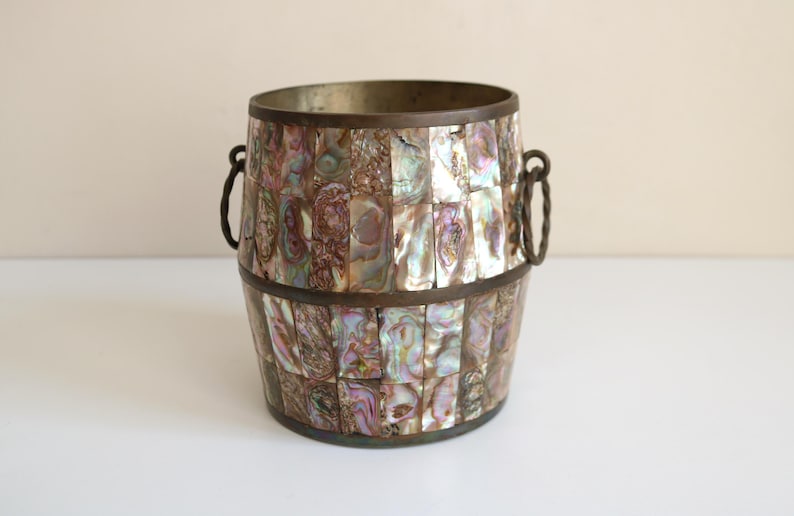May include: A decorative barrel-shaped container with a mosaic of iridescent abalone shell pieces. The container has a metal rim and handles, and it sits on a white surface. The colors include shades of purple, green, and brown.