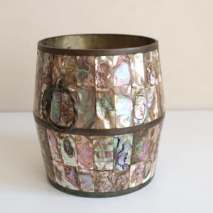 May include: A decorative barrel-shaped container with a mosaic of iridescent abalone shell pieces. The container has a dark metal rim and a metal handle. The colors include shades of pink, blue, and green.