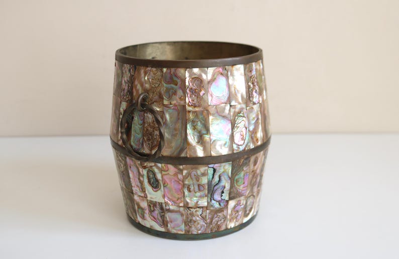 May include: A decorative barrel-shaped container with a mosaic of iridescent abalone shell pieces. The container has a dark metal rim and a metal handle. The colors include shades of pink, blue, and green.