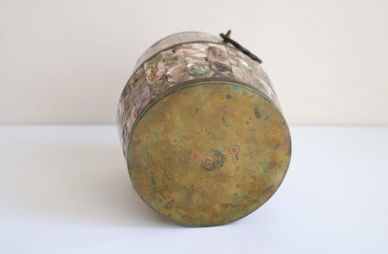 May include: A vintage barrel-shaped container with a brass base and lid. The body is adorned with a mosaic of iridescent, rectangular tiles in shades of pink, white, and gray. A dark metal clasp secures the lid.