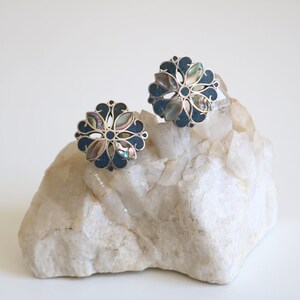 May include: A pair of silver earrings with a floral design. The earrings are made of silver and have a dark blue enamel background with iridescent abalone shell accents.