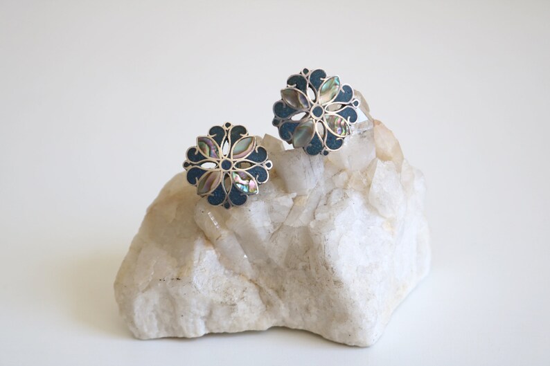 May include: A pair of silver earrings with a floral design. The earrings are made of silver and have a dark blue enamel background with iridescent abalone shell accents.