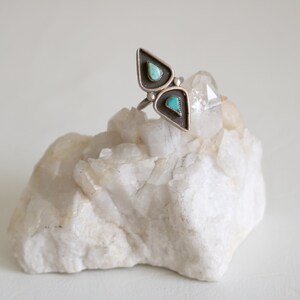 May include: A silver ring with two turquoise stones set in a triangular shape. The ring is resting on a white crystal.