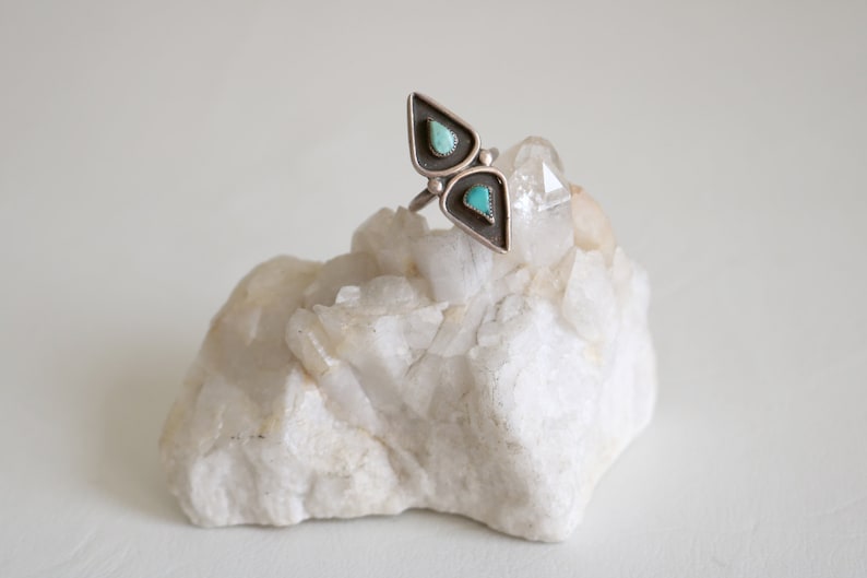 May include: A silver ring with two turquoise stones set in a triangular shape. The ring is resting on a white crystal.