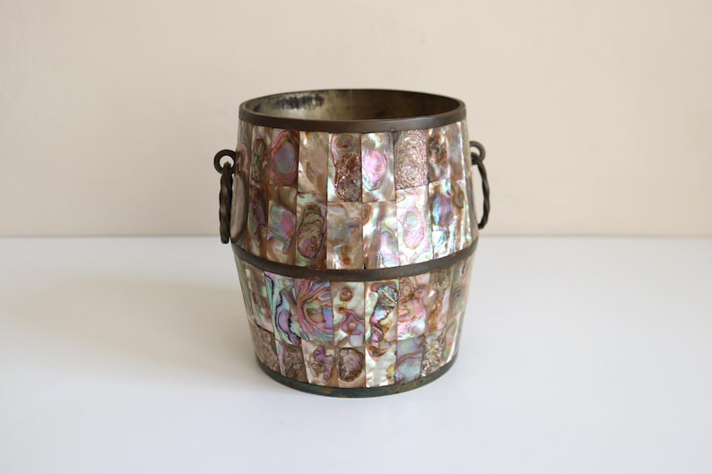 May include: A decorative barrel-shaped container with a mosaic design made of iridescent shell pieces. The container has a bronze-colored metal rim and handles. The shell pieces display a range of colors, including pink, purple, and green.