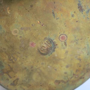 May include: Close-up of the bottom of a brass vessel with a weathered, aged appearance. The surface is a mix of gold and green hues, with small, reddish-brown spots. The text "HECHO EN MEXICO" is stamped on the bottom.