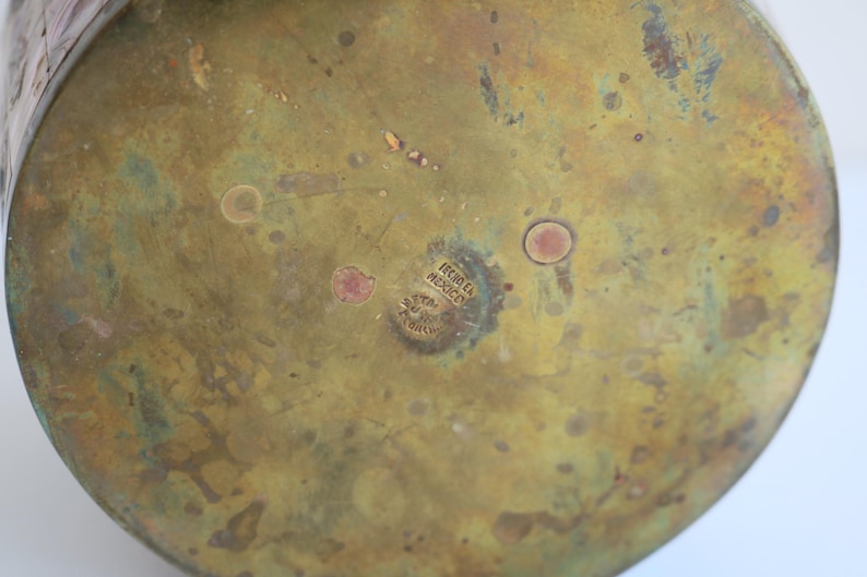May include: Close-up of the bottom of a brass vessel with a weathered, aged appearance. The surface is a mix of gold and green hues, with small, reddish-brown spots. The text "HECHO EN MEXICO" is stamped on the bottom.